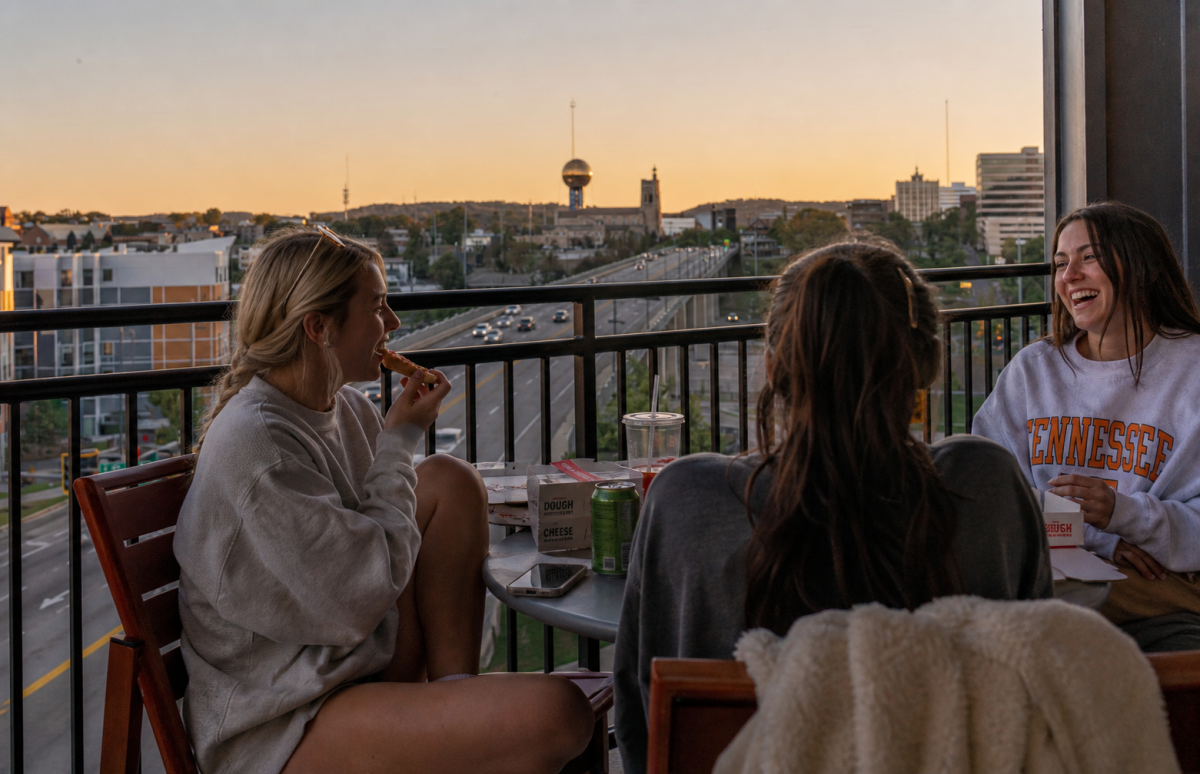 Three friends eat pizza and laugh on a balcony overlooking Knoxville at sunset