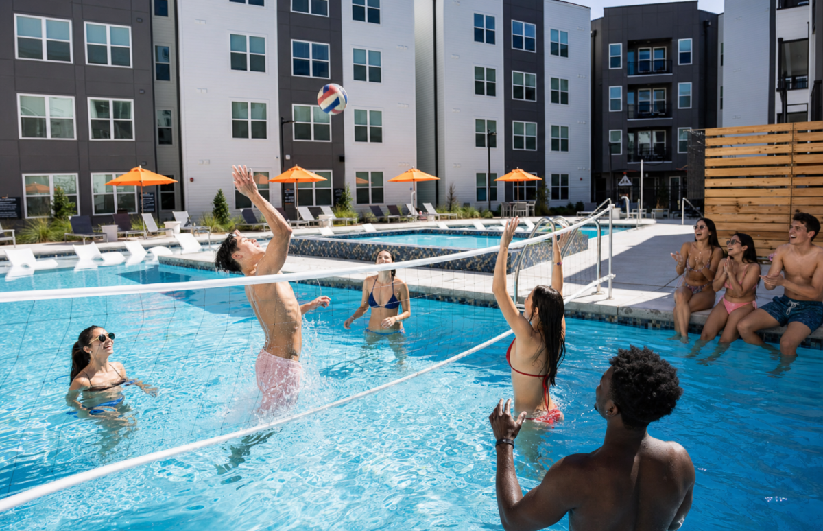 Friends play volleyball in an apartment pool while others cheer from the poolside
